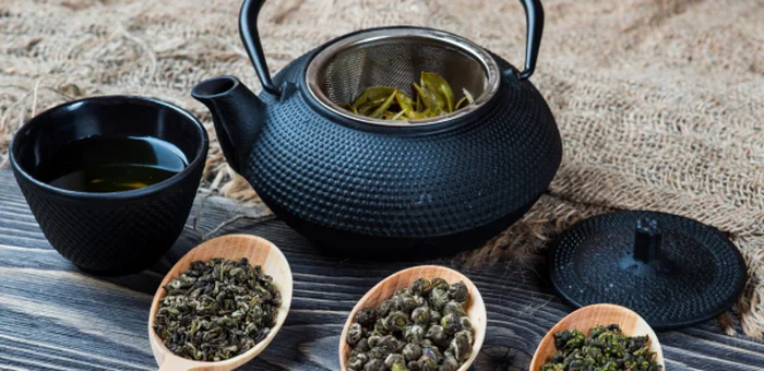 A cast-iron teapot with a cup of herbal tea and three wooden spoons of dried tea leaves.